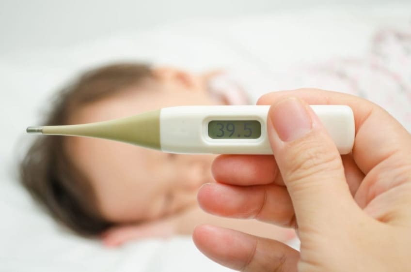Mother holding a thermometer in the foreground with a sick child sleeping in the background