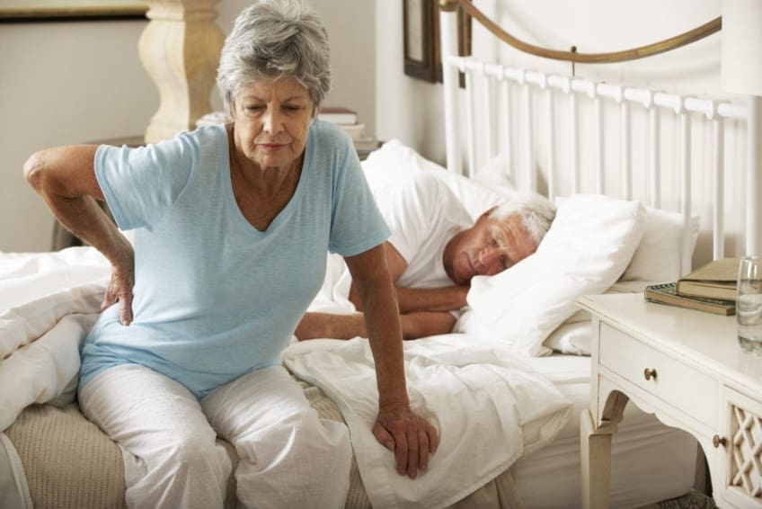 older woman sits on bed with her hand on her hip in pain