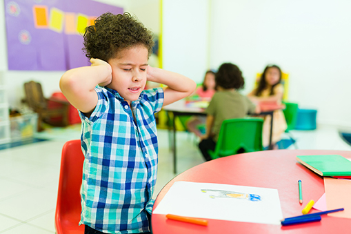 Upset autistic little boy covering his ears and feeling distressed and overwhelmed by the loud noises in preschool