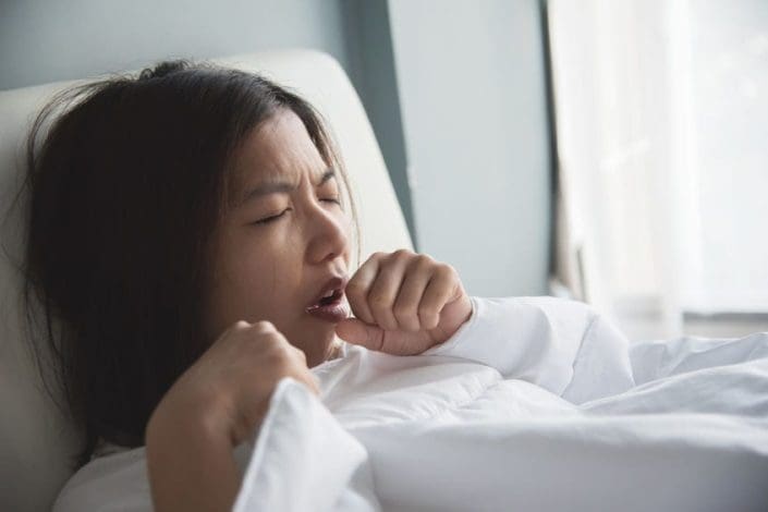 A woman is coughing on her bed. Finding the best cough suppressant can be a challenge.