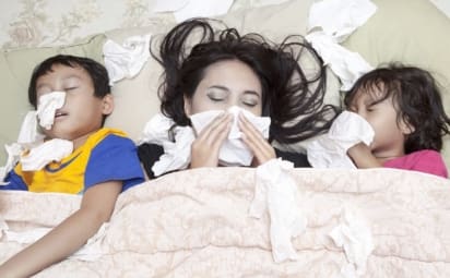 A mother and two young children lie in bed blowing their noses. Flu symptoms can sometimes be mistaken for symptoms of the common cold and vice versa. Both are contagious respiratory illnesses.