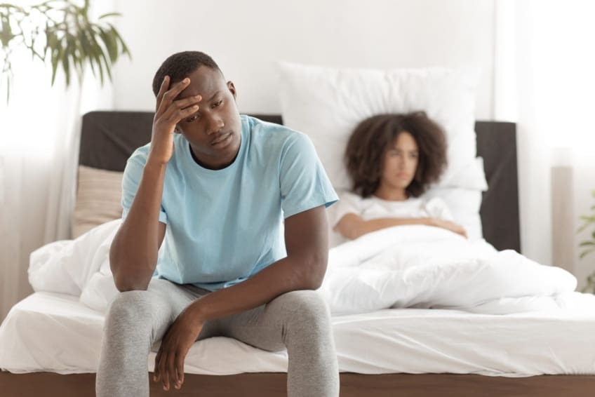 Male struggling with premature ejaculation sits on edge of bed with girlfriend in the background.