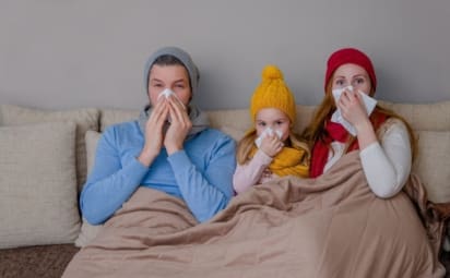 A family suffering from the flu holds tissues up to their noses while sitting on a couch.