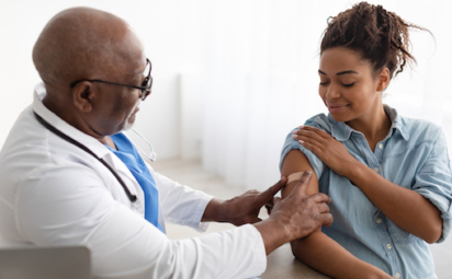 A doctor in a white lab coat with a stethoscope places a Band-Aid on the upper arm of a patient who just received a flu vaccine.