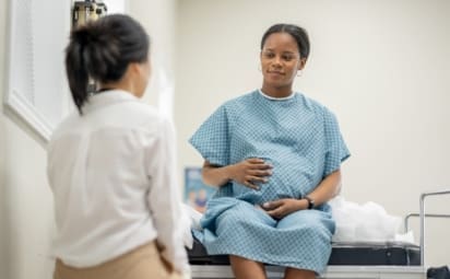 A young pregnant woman sit up on an exam table in her doctors office during a routine prenatal check-up. She wearing a medical gown and her doctor is seated across from her as the two talk.