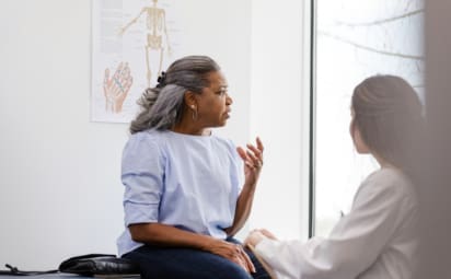 A woman, appearing worried, is seated on an exam table while talking with her doctor.
